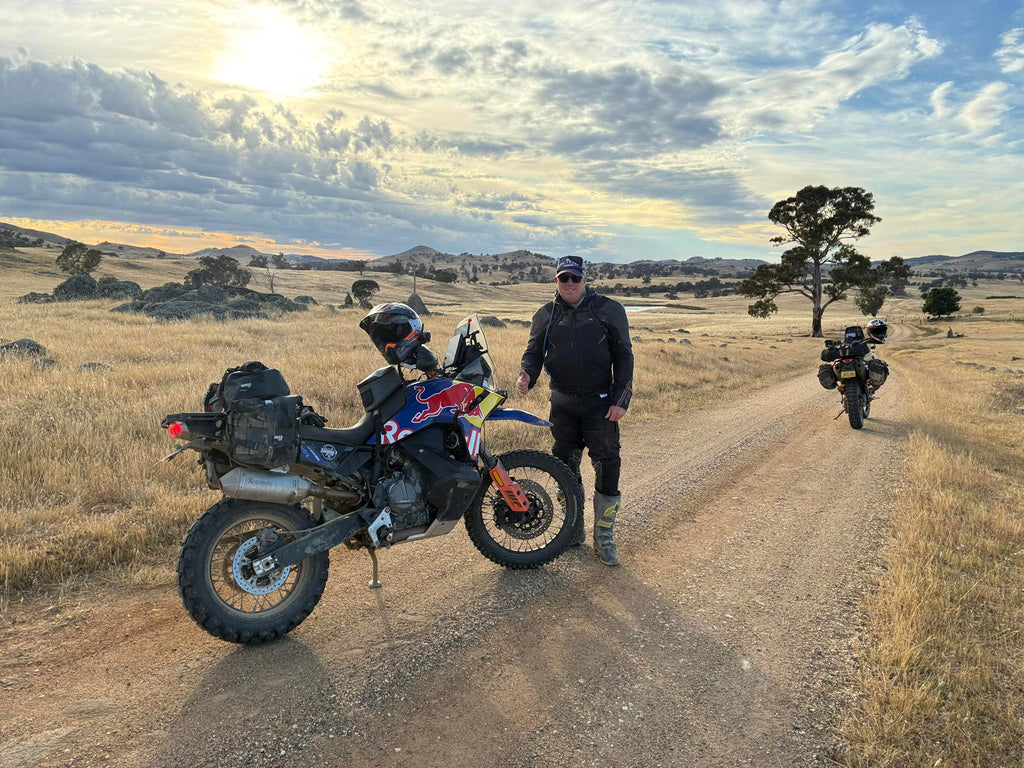 Man standing next to a motorcycle on a dirt road with another motorcycle in the background under a cloudy sky.