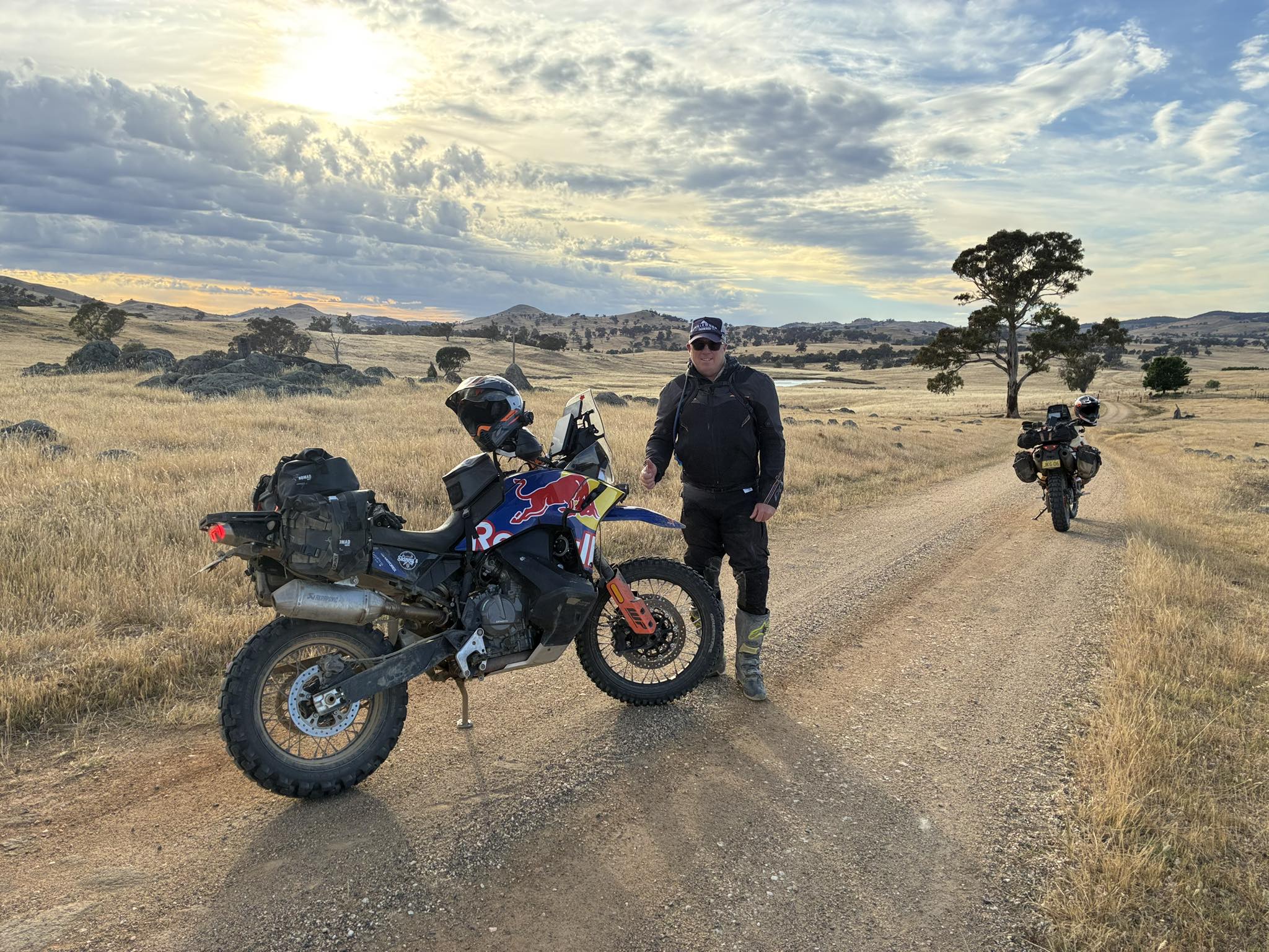 Man standing next to a motorcycle on a dirt road with another motorcycle in the background under a cloudy sky.